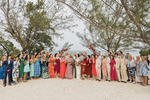 Large multicultural wedding group photo on a private island resort, with colorful attire and tropical trees framing the ocean backdrop.